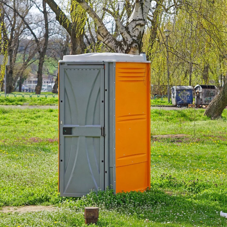 A portable toilet with a gray door and an orange side, situated in a grassy area with trees in the background.