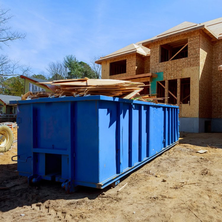 A large blue dumpster filled with construction debris, including wooden planks, is situated next to a partially constructed building. The building has exposed framing and windows, with a clear blue sky in the background.