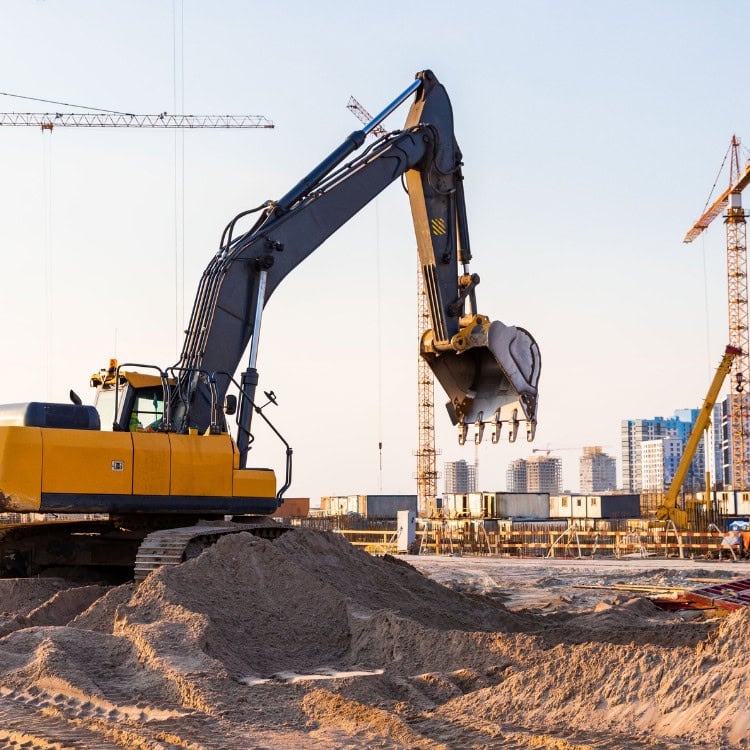 An excavator is positioned on a construction site, with its arm extended and bucket hovering above a pile of sand. In the background, there are construction cranes and buildings under development. The scene is set against a clear sky during the late afternoon.