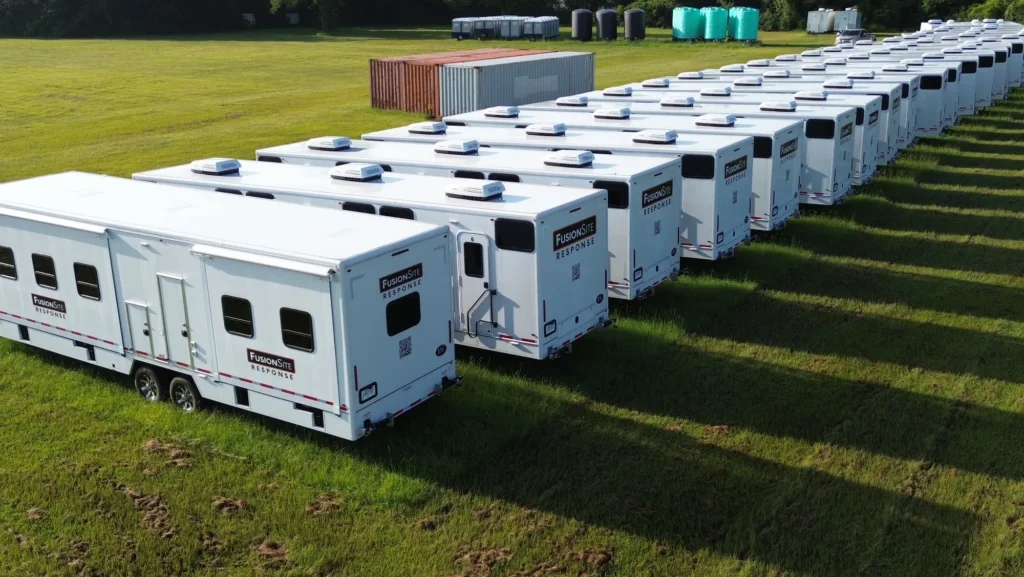 A row of white mobile restroom trailers for FusionSite Response is staged for deployment on a large paved lot under overcast skies, with support trucks and equipment positioned nearby.