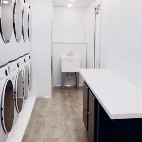 A bright mobile laundry trailer interior featuring stacked washer-dryers, a countertop with storage, a utility sink, and wood-style flooring beneath white walls and recessed ceiling lights.