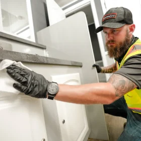 A worker wearing a safety vest and gloves is cleaning a countertop inside a portable restroom, with a mirror and door visible in the background.
