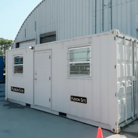 A white portable restroom unit with a "Fusion Site" sign is positioned beside a blue shipping container on a paved area outside a large metal building under a clear blue sky.
