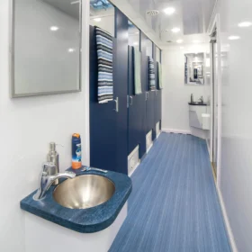 A clean restroom trailer interior featuring blue stall doors with hanging towels, a stainless-steel sink with soap and toiletries, mirrors, and blue-striped flooring under bright ceiling lights.