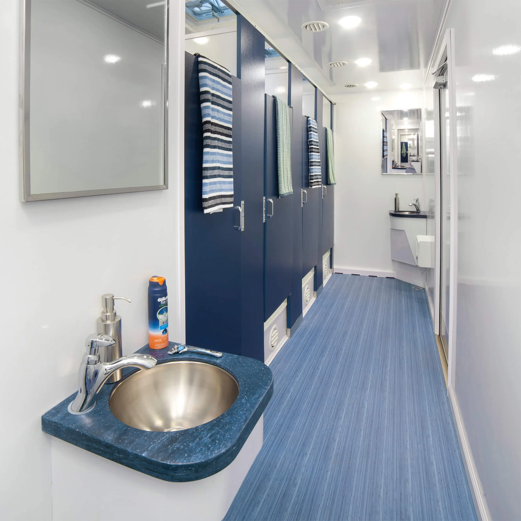 A clean restroom trailer interior featuring blue stall doors with hanging towels, a stainless-steel sink with soap and toiletries, mirrors, and blue-striped flooring under bright ceiling lights.