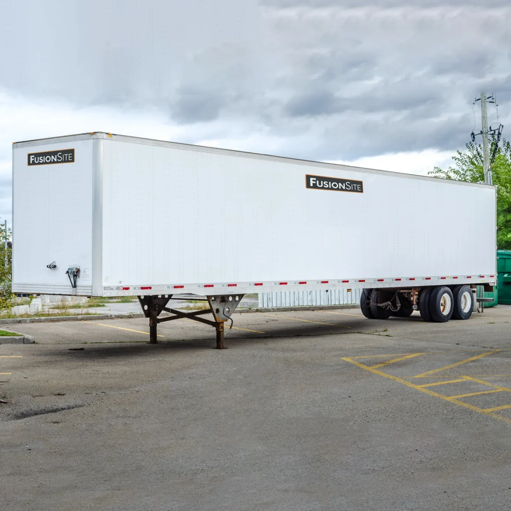 A large white trailer with the logo "FusionSite" is parked on a concrete lot, surrounded by overgrown grass and a green dumpster, under a cloudy sky.