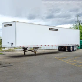 A large white trailer with the logo "FusionSite" is parked on a concrete lot, surrounded by overgrown grass and a green dumpster, under a cloudy sky.