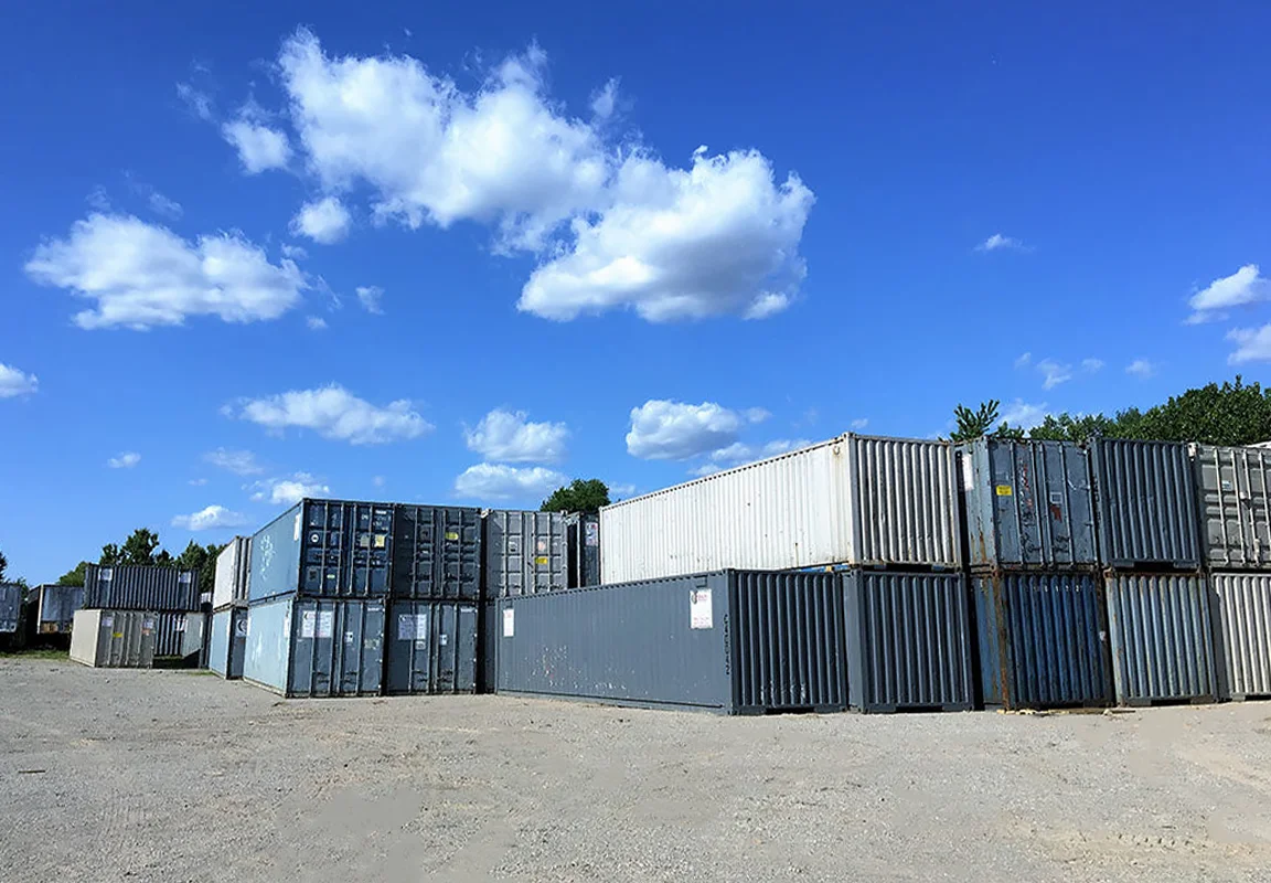A large open lot with stacks of 40-foot shipping containers in shades of gray and blue under a bright blue sky with scattered white clouds.
