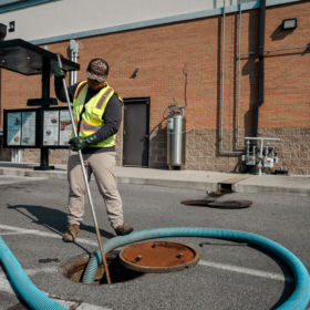 A worker in a reflective vest is using a blue hose to service a manhole in a parking lot beside a brick building, with utility pipes visible on the wall in the background.