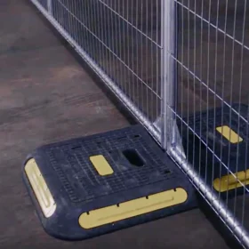 A close-up of a temporary fence base with a heavy-duty black and yellow Big Foot weight support block securing the metal fencing post.