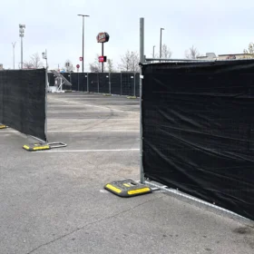 A series of black fencing panels is set up in a parking lot, with yellow weights securing the bases, under a cloudy sky and near a fast-food restaurant sign.