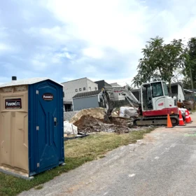 A blue and beige portable restroom is positioned on a gravel path next to a construction site, with a small excavator and piles of dirt nearby under a cloudy sky.