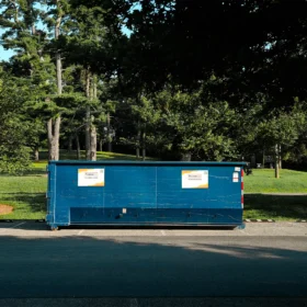 A blue dumpster with company signage is parked on a paved area beside a grassy field and trees under a clear blue sky.