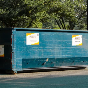A large blue dumpster with company signage is parked on a gravel surface beside a grassy area, surrounded by trees on a sunny day.