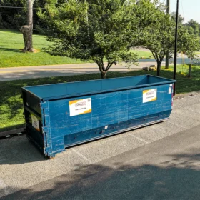 A large blue dumpster with company branding is positioned on a paved parking lot, surrounded by grassy areas and trees, with a road and a fountain visible in the background.