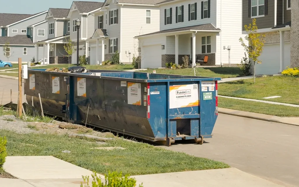 A large blue dumpster is positioned on a paved street in front of newly constructed homes, with neatly manicured lawns and clear blue skies in the background.