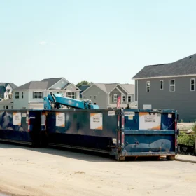 Two large blue dumpsters are positioned on a dirt road in front of several newly constructed houses, with a construction crane visible in the background under a clear blue sky.