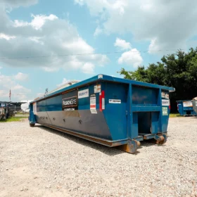 A row of blue dumpsters is parked on a gravel lot under a partly cloudy sky, with additional dumpsters visible in the background near a tree line.