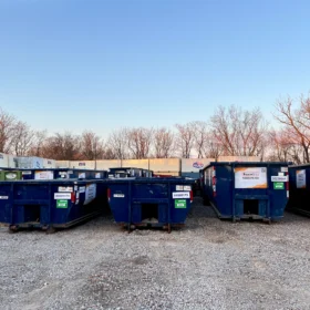 A row of blue dumpsters with various labels is arranged on a gravel lot, surrounded by bare trees and a clear sky in the background.