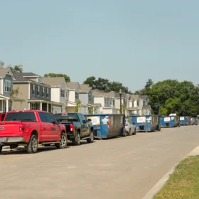 A line of blue dumpsters is parked along a newly paved street in a residential neighborhood, with modern houses and green trees visible in the background under a clear blue sky.