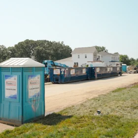 A blue portable restroom stands beside a row of large dumpsters on a construction site, with a residential building and trees in the background under a clear blue sky.