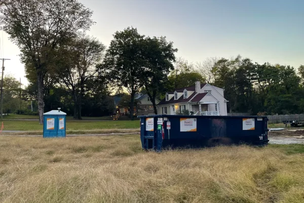 A blue portable restroom and a large dark dumpster are positioned on a grassy area near a residential home, surrounded by trees and power lines during dusk.