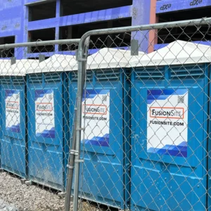 A row of blue portable restrooms is secured behind a chain-link fence at a construction site, with a partially constructed building visible in the background on a cloudy day.