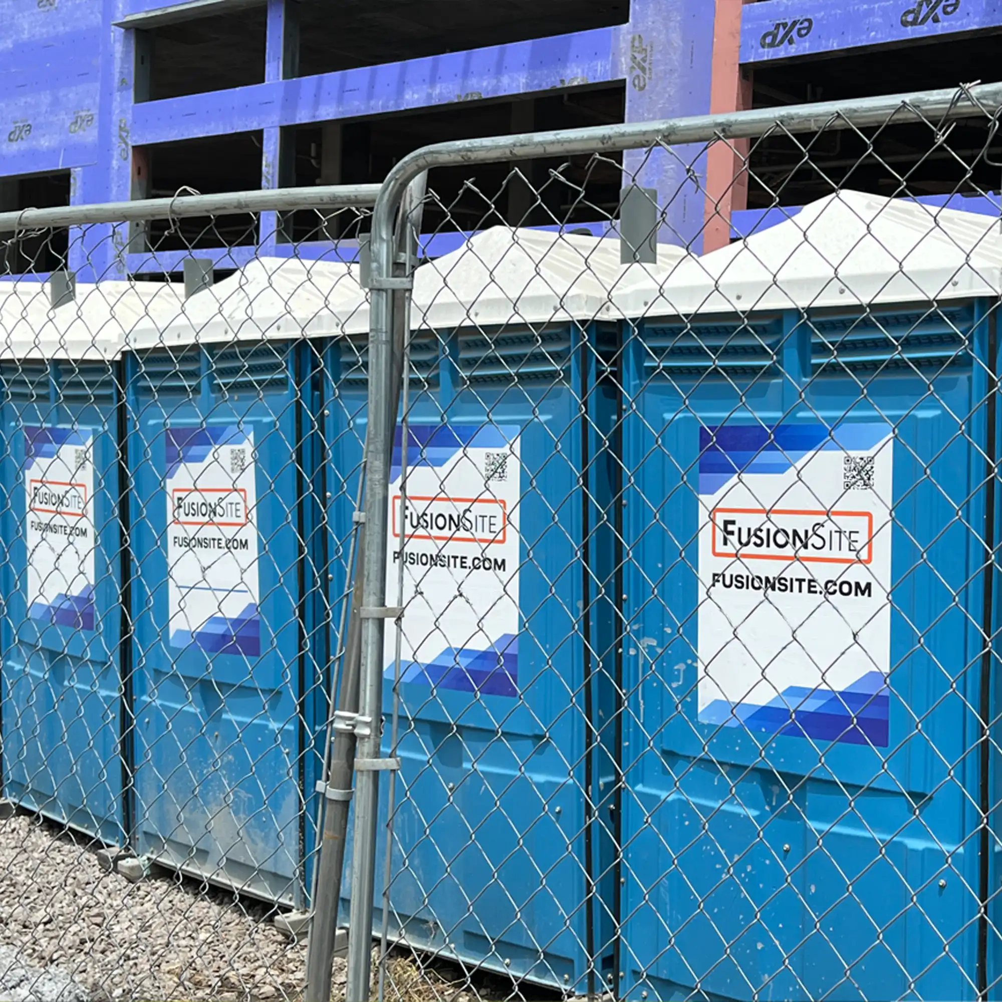A row of blue portable restrooms is secured behind a chain-link fence at a construction site, with a partially constructed building visible in the background on a cloudy day.