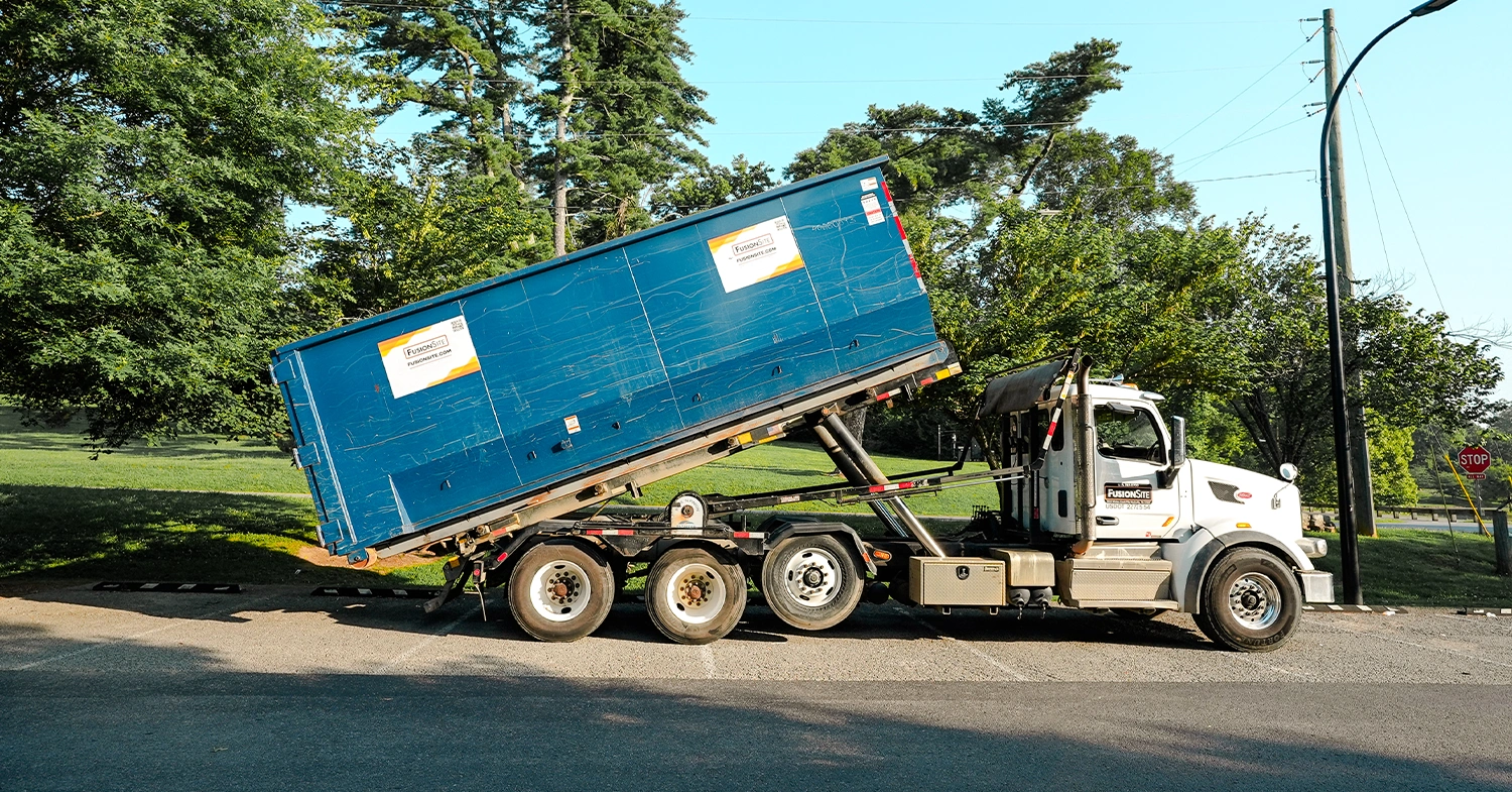 A blue roll-off dumpster is tilted on the back of a white truck, parked on a paved road near a grassy area and a stop sign under a clear blue sky.