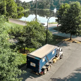 A blue sanitation truck with a covered bed is parked in a shaded area beside a grassy field, near a calm pond and surrounded by tall trees on a sunny day.