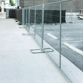 A chain-link fence encloses a construction area beside a modern building, with a portable restroom visible in the background and a paved walkway leading towards the entrance.