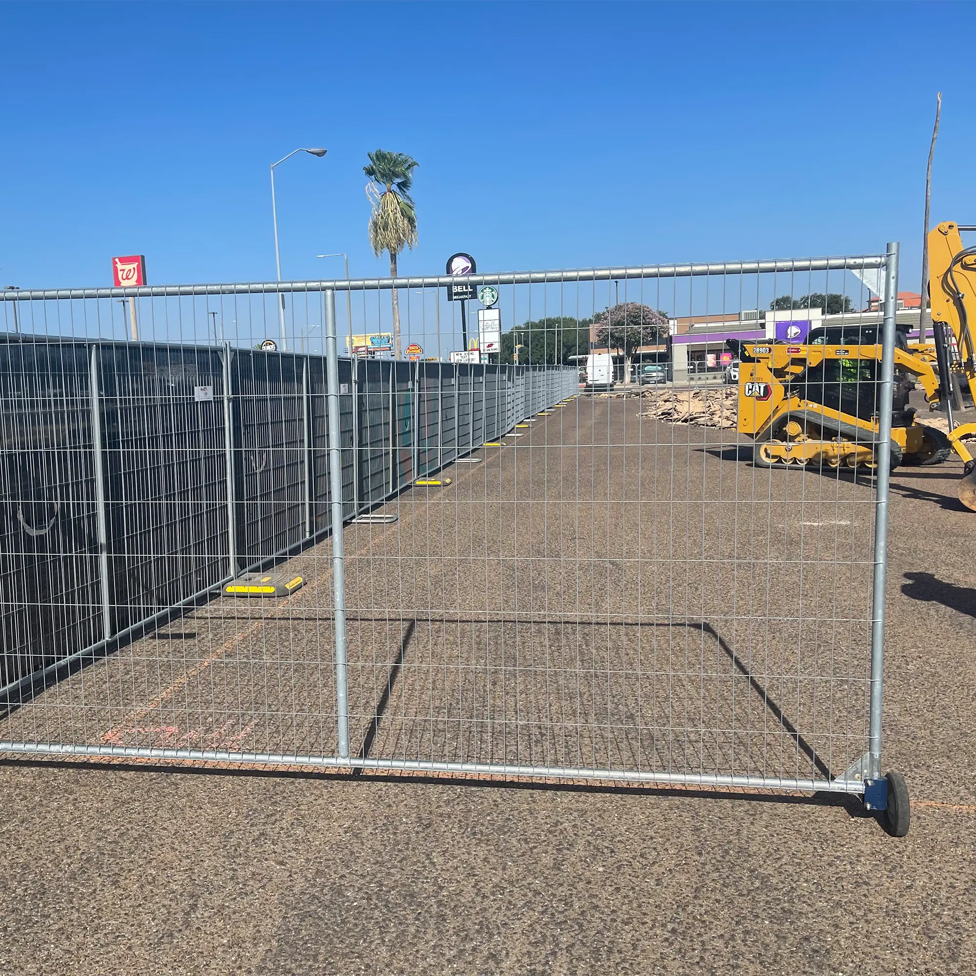 A chain-link construction fence encloses a gravel area with portable restrooms and a yellow excavator, set against a clear blue sky and nearby commercial buildings.