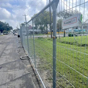 A series of black fencing panels is set up in a parking lot, with yellow weights securing the bases, under a cloudy sky and near a fast-food restaurant sign.