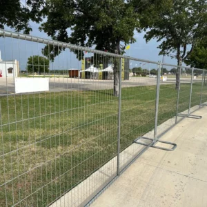 A chain-link fence encloses a grassy area beside a paved driveway, with a large sign for a portable restroom service visible in the background under a partly cloudy sky.