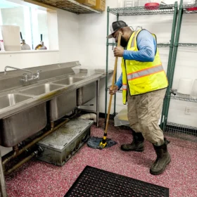 A worker in a high-visibility vest is mopping the floor in a commercial kitchen, with stainless steel sinks and shelves filled with kitchen supplies in the background.