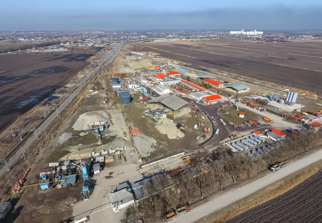 An aerial view of a large construction site features multiple storage containers, machinery, and vehicles, with a backdrop of open fields and a distant industrial area under a clear blue sky.