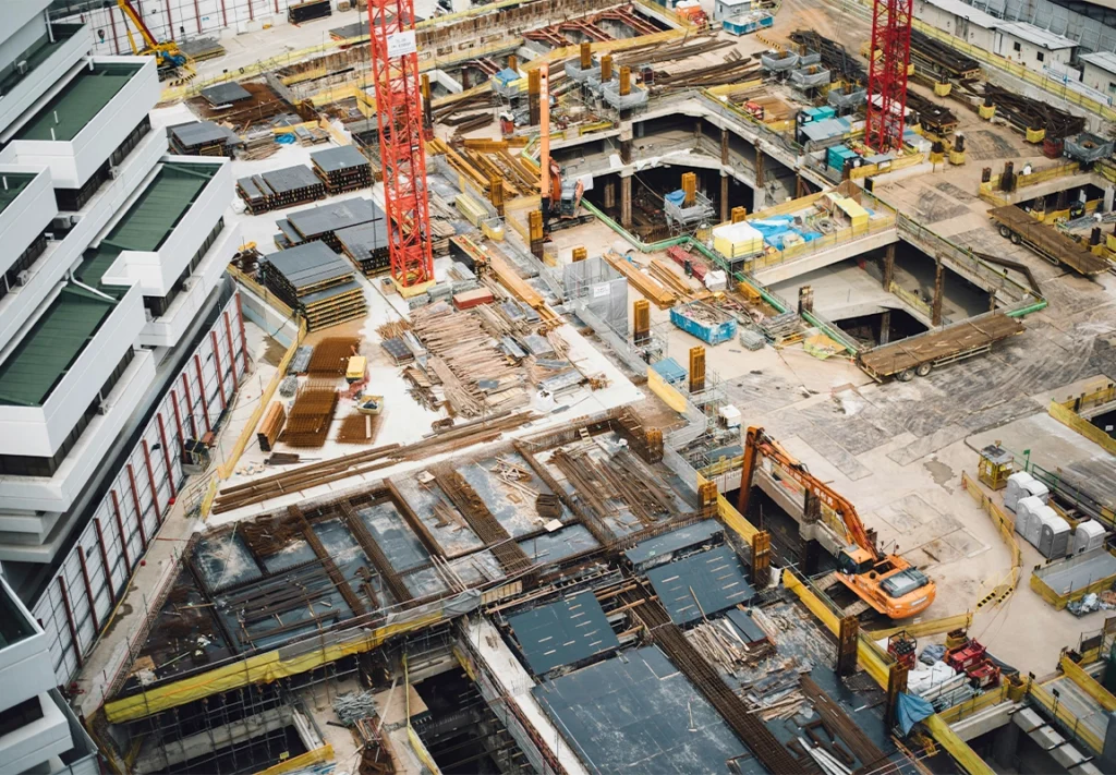 A construction site bustling with activity features heavy machinery, stacks of building materials, and several portable restrooms positioned near the edge of a partially excavated area under a cloudy sky.