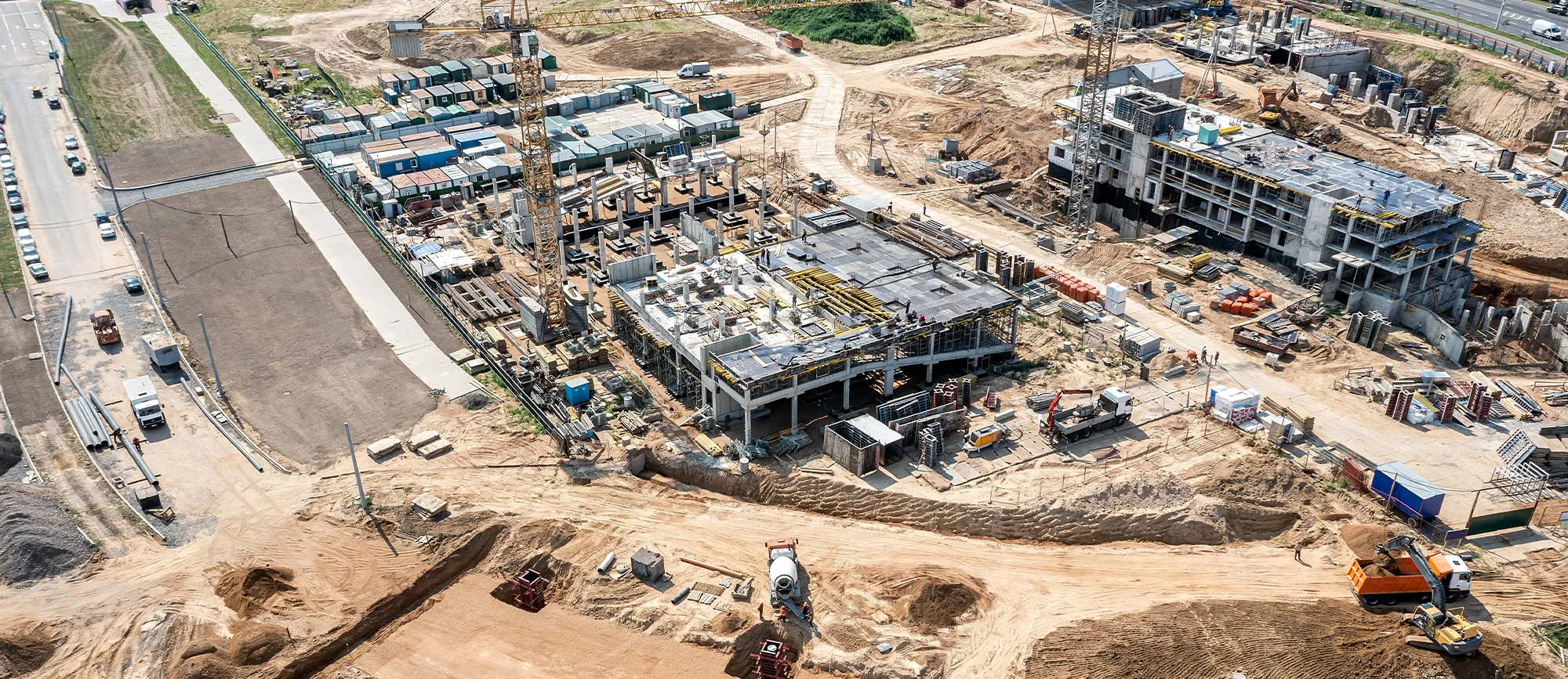 A construction site features several large machinery vehicles, stacks of building materials, and a partially constructed building surrounded by dirt and gravel, with a road visible in the background.