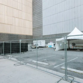 A construction site is enclosed by metal fencing, featuring a white tent and two service trailers parked on a paved area beside a large building with a wooden facade.