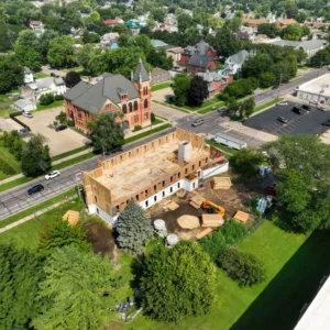 A construction site with a partially built wooden structure is surrounded by trees and grass, with construction equipment and materials scattered on the ground, located near a street and residential buildings.