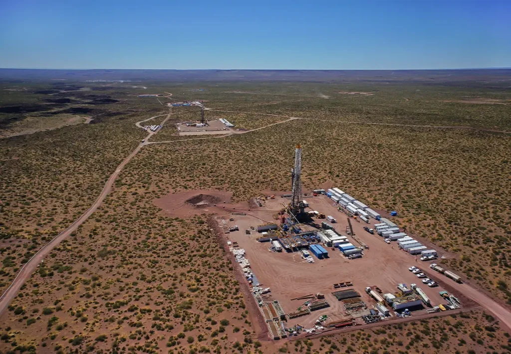 A large drilling rig is set up in a remote area surrounded by sparse vegetation, with multiple service vehicles and storage containers arranged on a flat, sandy surface under a clear blue sky.