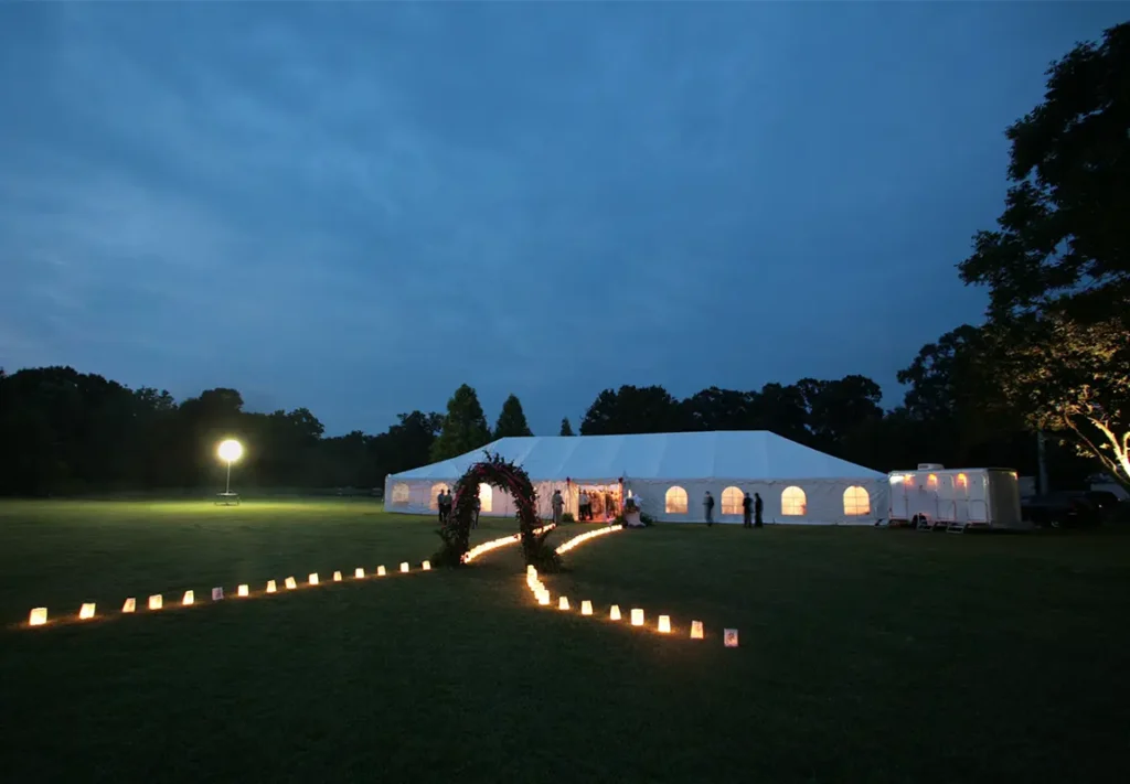 A large white event tent is set up on a grassy field at dusk, illuminated by lanterns lining a pathway, with people mingling near the entrance.
