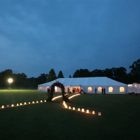 A large white event tent is set up on a grassy field at dusk, illuminated by lanterns lining a pathway, with people mingling near the entrance.