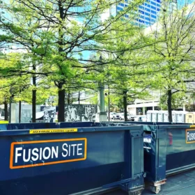 A large blue dumpster labeled "Fusion Site" is positioned on a concrete surface surrounded by green trees, with several portable restrooms visible in the background near a modern building.