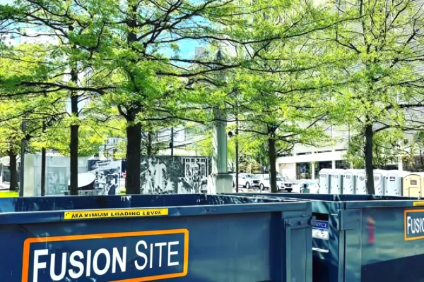 A large blue dumpster labeled "Fusion Site" is positioned on a concrete surface surrounded by green trees, with several portable restrooms visible in the background near a modern building.