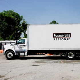 A white delivery truck with "FusionSite Response" branding is parked on a gravel lot, with several portable restrooms visible in the background under a clear blue sky.