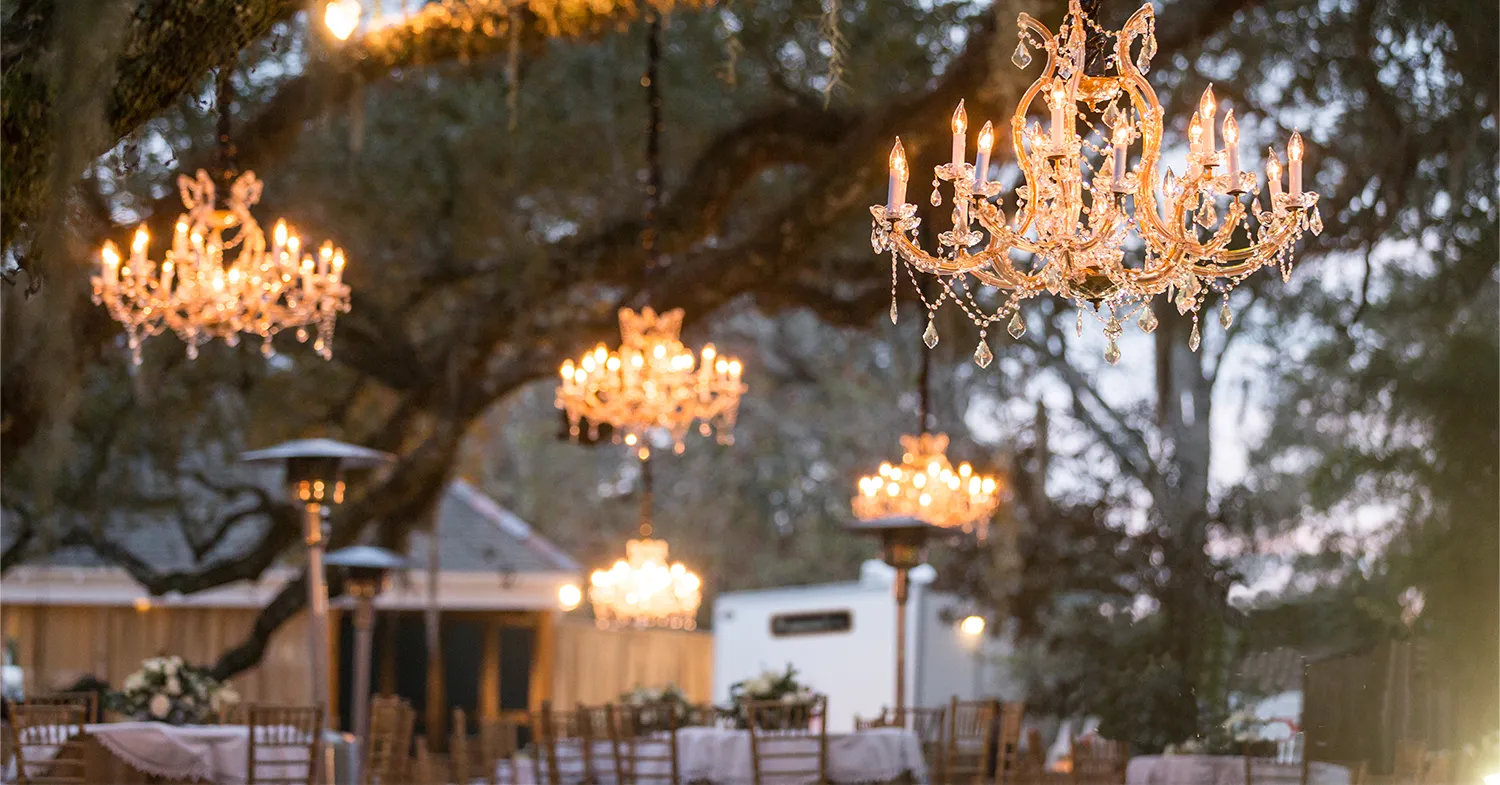 A white portable restroom trailer is positioned outdoors among elegantly arranged tables and hanging chandeliers, with trees and a wooden structure visible in the background during twilight.