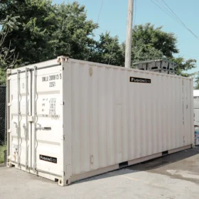 A large white shipping container is positioned on a gravel surface beside a chain-link fence, with portable restrooms visible in the background under a clear blue sky.