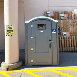 A gray portable restroom is positioned beside a beige column and a wooden fence, with yellow no-parking lines visible on the pavement and signage indicating fire lane restrictions.