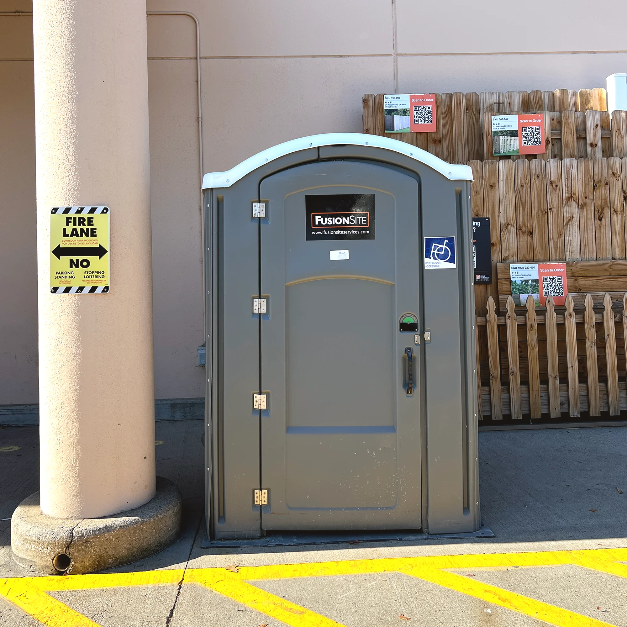 A gray portable restroom is positioned beside a beige column and a wooden fence, with yellow no-parking lines visible on the pavement and signage indicating fire lane restrictions.